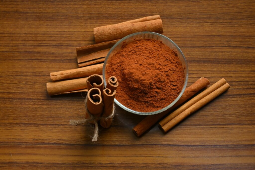 Pote de vidro com canela em pó, com pedaços de canela em pau ao redor em cima de mesa de madeira