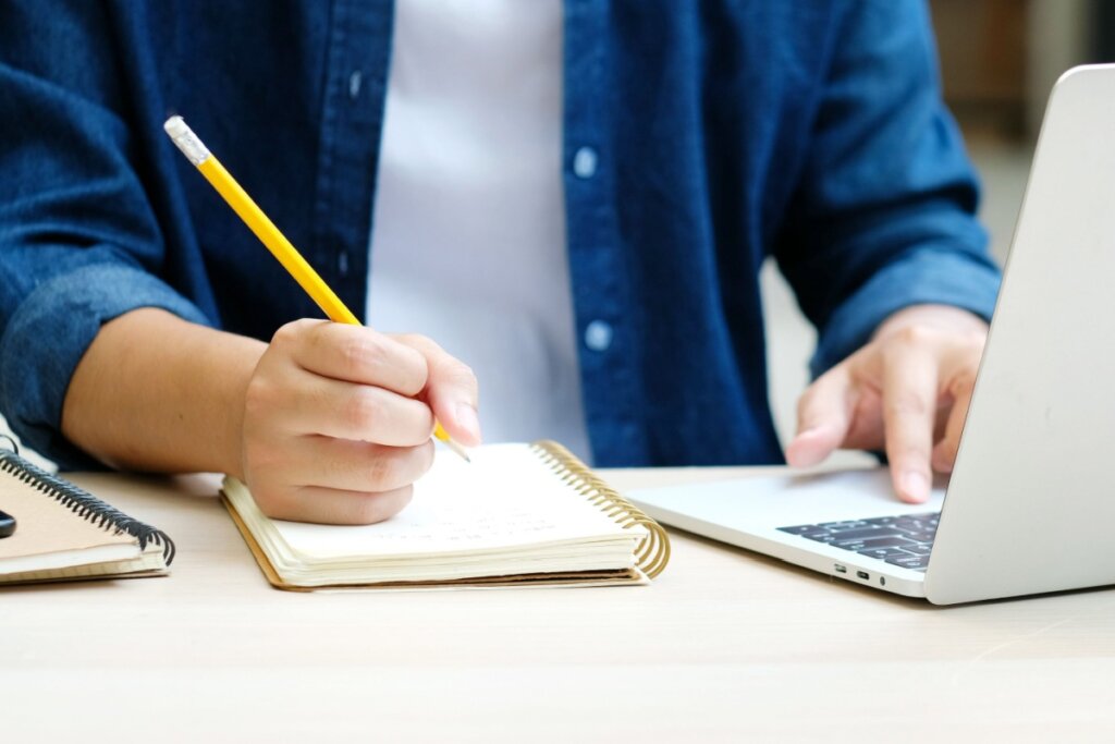 Mão anotando algo com um lápis em um caderno. Notebook cinza do lado 