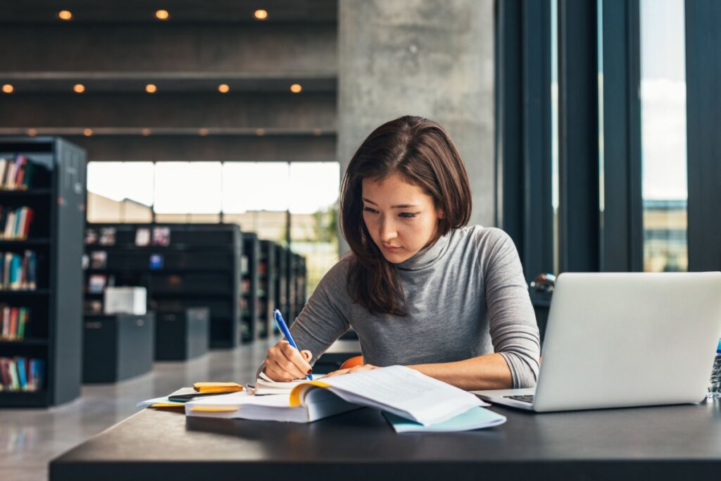 Uma jovem de cabelos castanhos e blusa cinza de gola alta estuda concentrada em uma biblioteca moderna. Ela está sentada à mesa, escrevendo em um caderno com uma caneta azul, cercada por livros abertos e um notebook prateado. Ao fundo, veem-se estantes de livros pretas e uma arquitetura industrial com colunas de concreto e grandes janelas que iluminam o ambiente.
