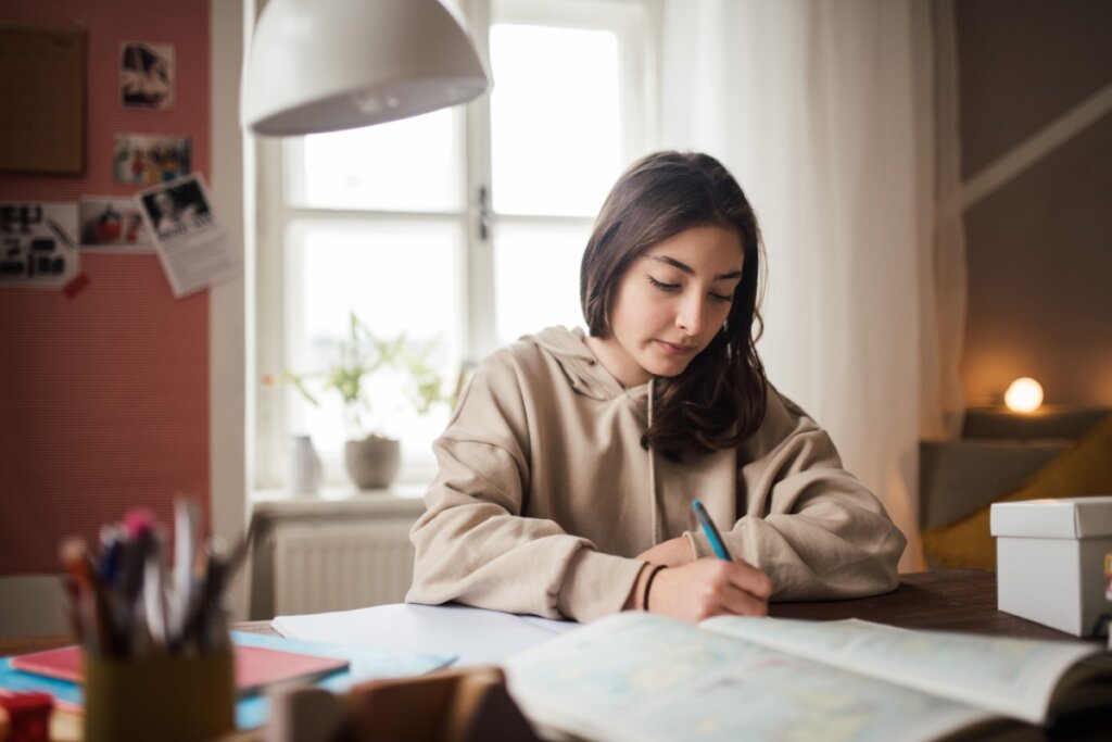Jovem com o cabelo curto e liso, usando blusa de moletom bege, estudando e escrevendo em papel