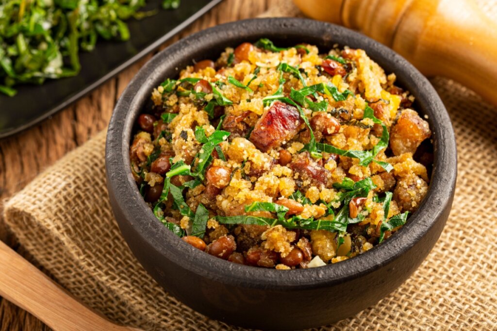 Feijão-tropeiro servido em tigela de cerâmica preta em cima de mesa de madeira