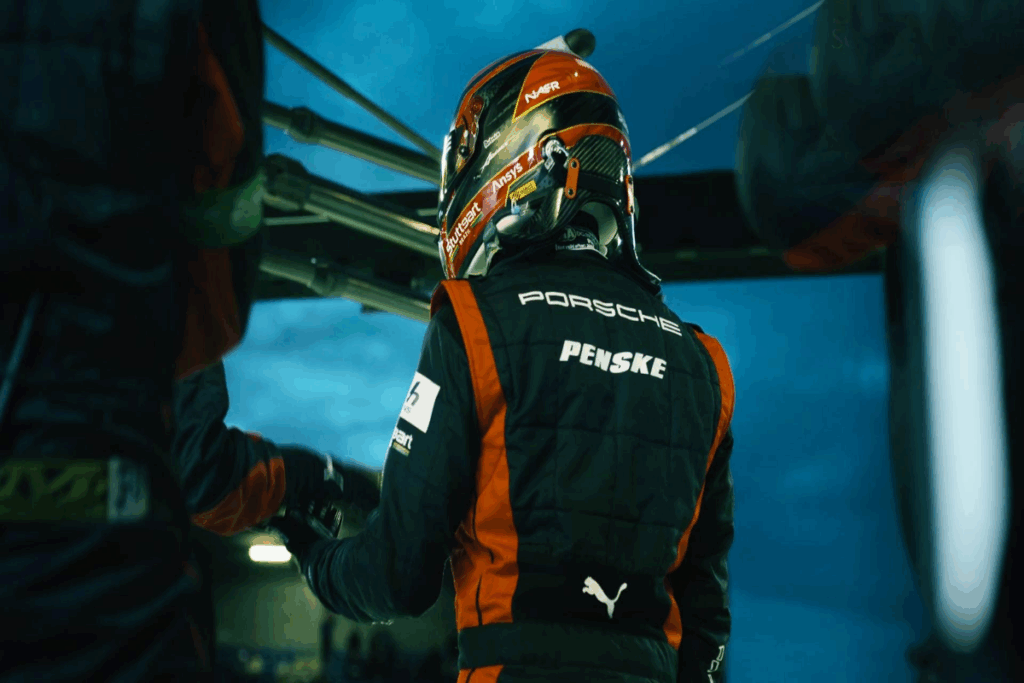 Homem usando roupa de piloto de carro preta com detalhes em vermelho e branco e usando capacete preto com detalhes em vermelho de costas