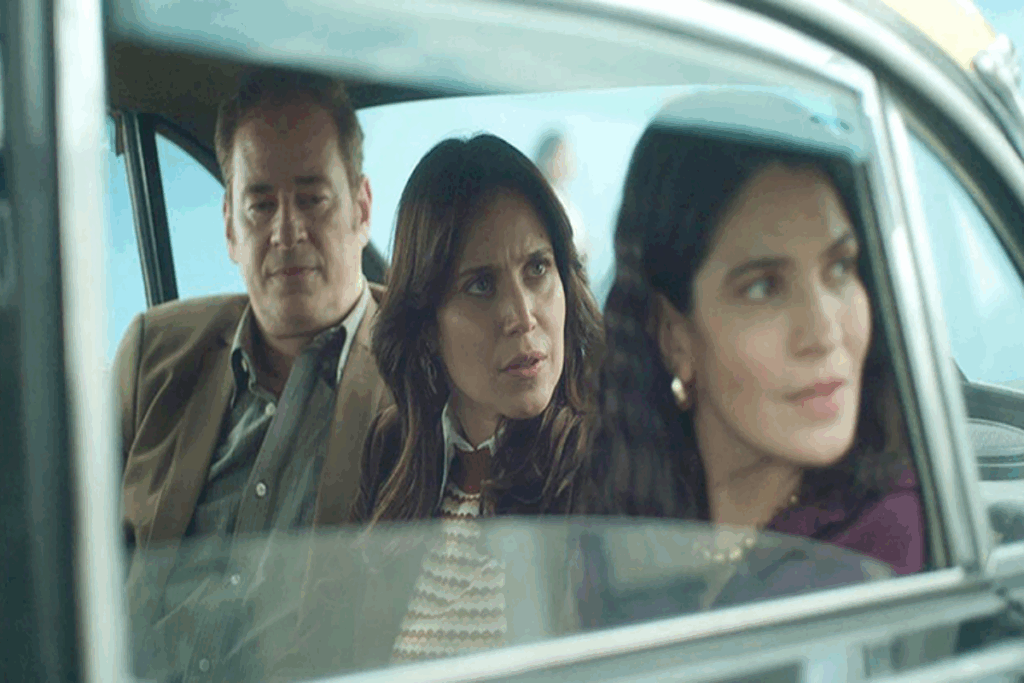 Imagem de duas mulheres com cabelo solto e usando roupas em tons de marrom, com homem com cabelo curto, usando camisa cinza e terno marrom sentados em banco de trás de carro