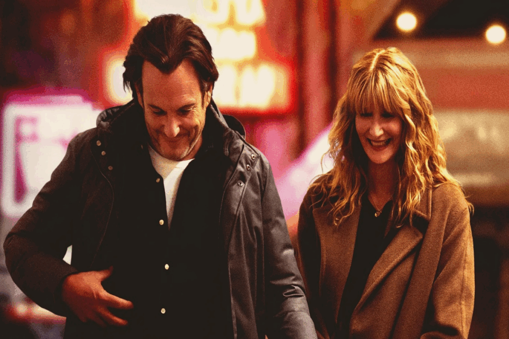 Homem com cabelo longo, usando camiseta branca e camisa preta e jaqueta escura andando ao lado de mulher com cabelo loiro usando camisa escura e casaco bege com luzes da rua ao fundo