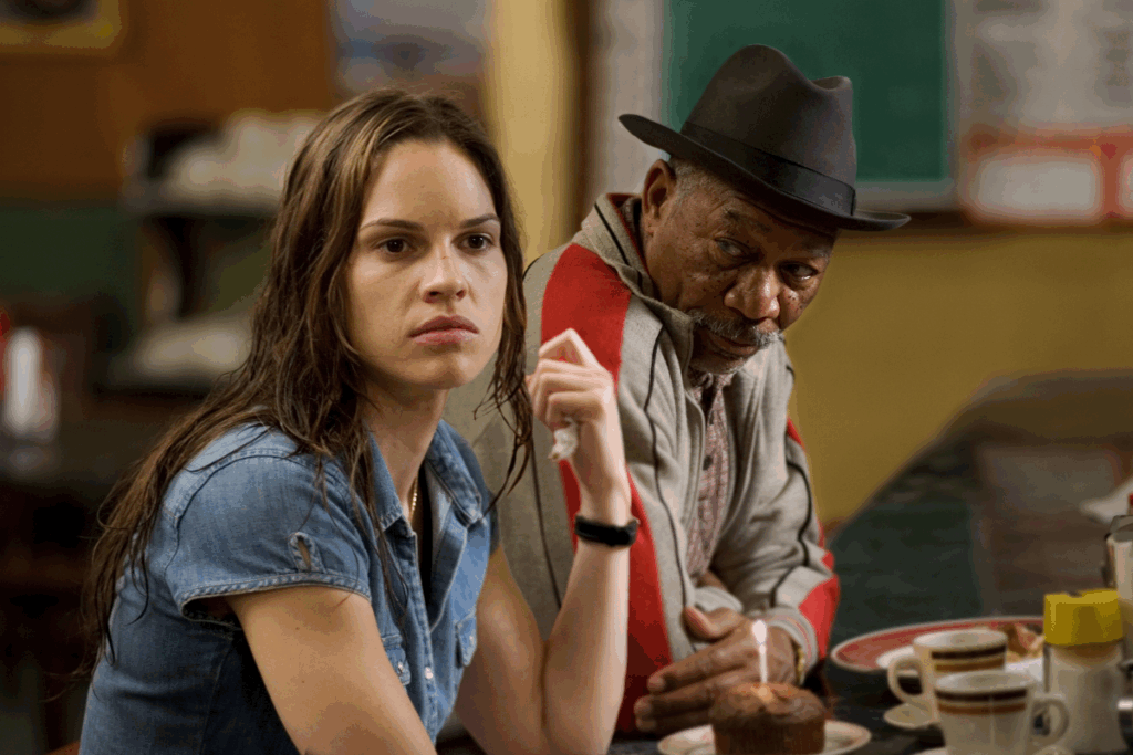 Imagem de uma jovem com o cabelo liso, solto e molhado, usando camiseta jeans e com um guardanapo na mão ao lado de um homem usando um chapéu preto e uma blusa de frio cinza com detalhes em vermelho e preto sentados em bancada de bar