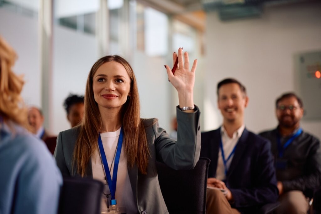 Mulher usando crachá e sorrindo, sentada em uma sala com uma das mãos levantadas 