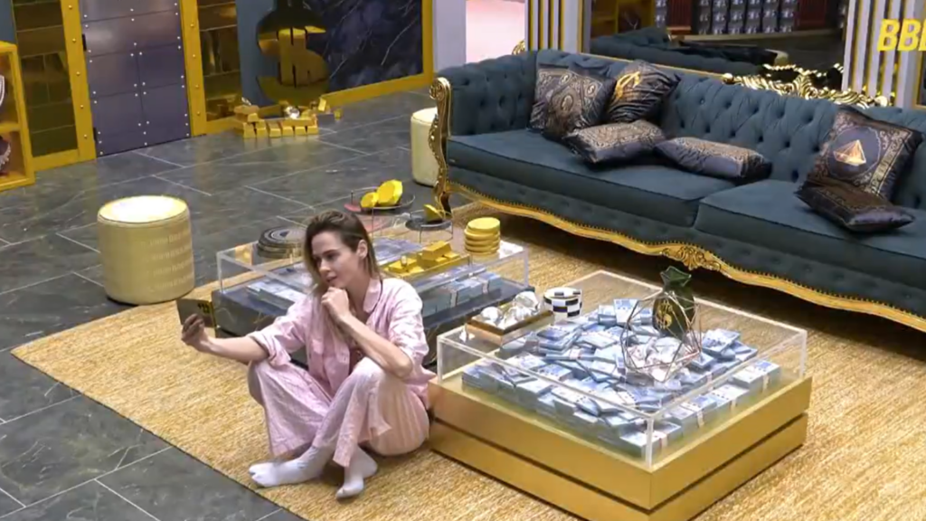 Ana Paula sentada no chão da sala do BBB enquanto grava um vídeo com um celular na horizontal