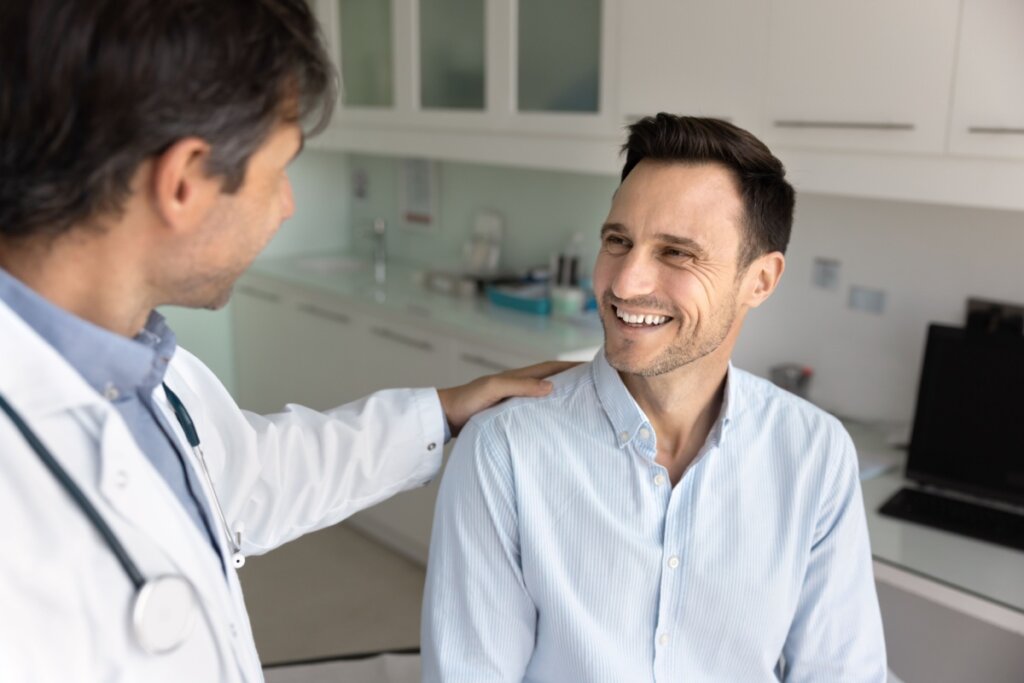 Homem com cabelo curto, e barba usando camisa de botões em consulta com médico que tem o cabelo curto, barba, está usando jaleco branco e com estetoscópio no pescoço