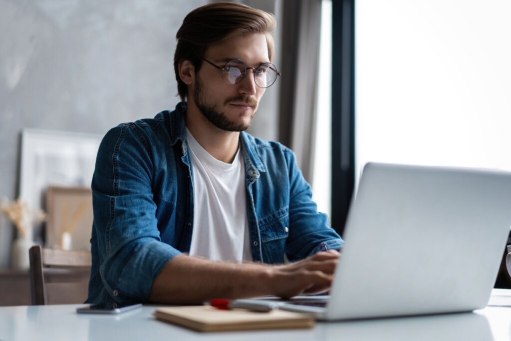 homem de camisa branca e jeans sentado e mexendo em notebook