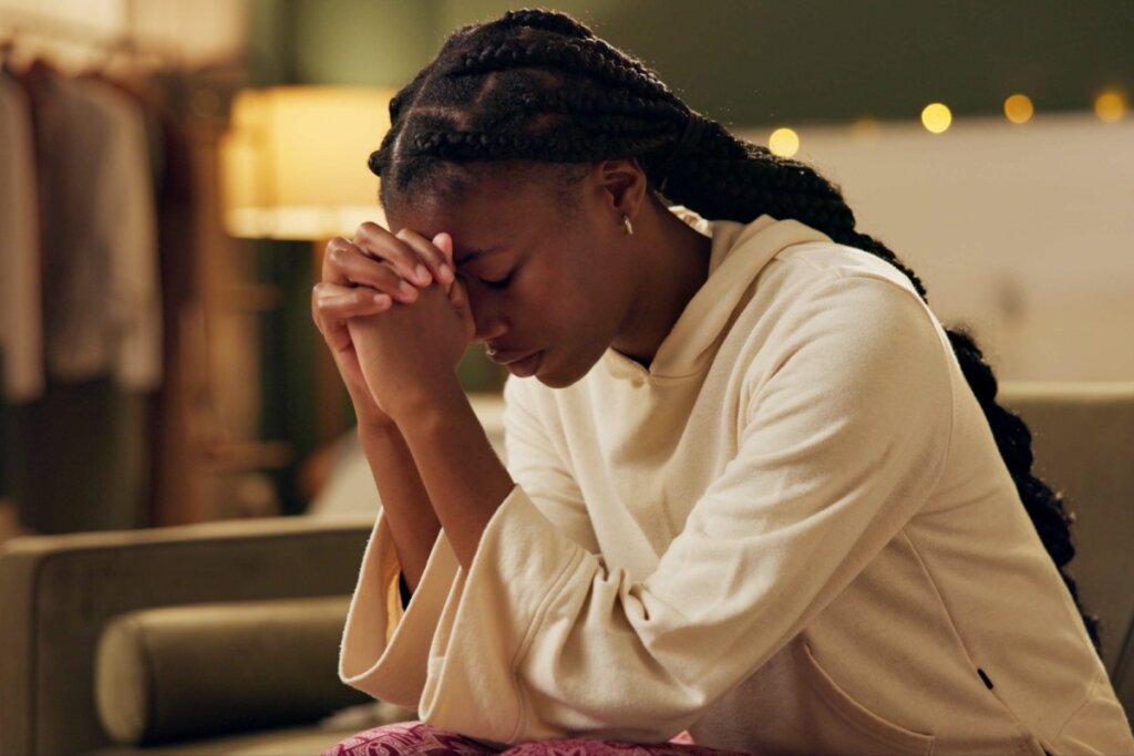 Jovem negra com o cabelo trançado e usando um casaco branco orando com as mãos unidas junto ao rosto 