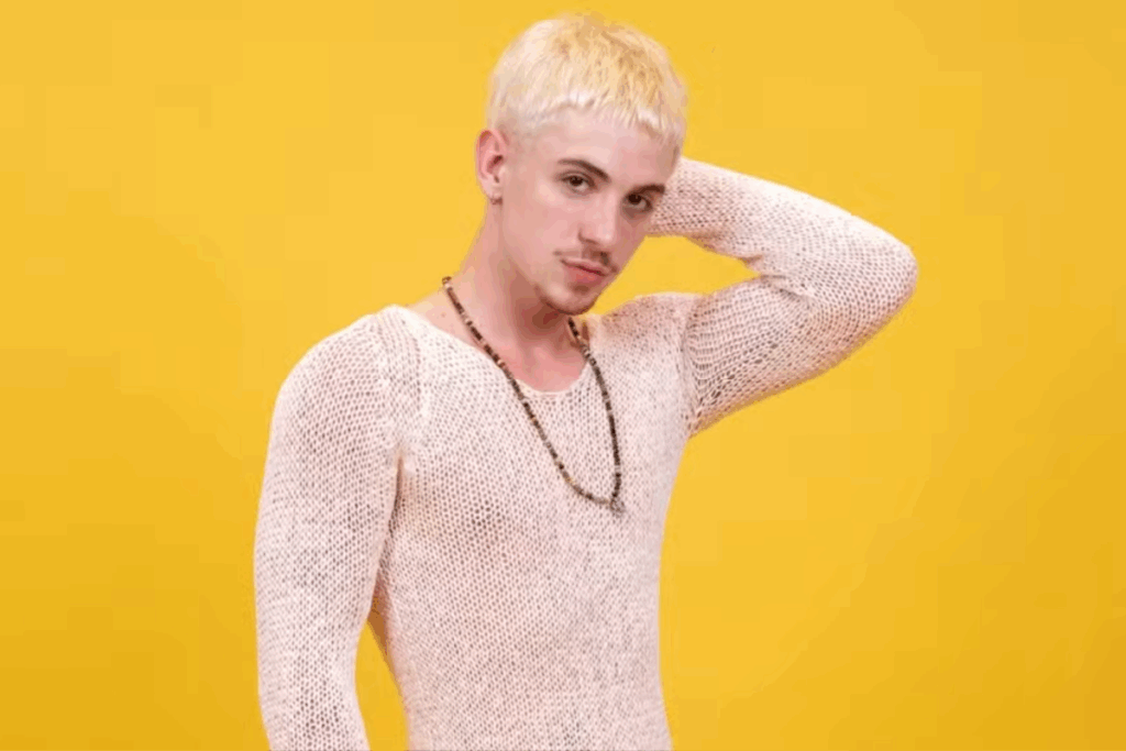 Homem jovem de cabelo loiro curto posando com expressão séria, usando blusa de tricô clara e colar, em fundo amarelo