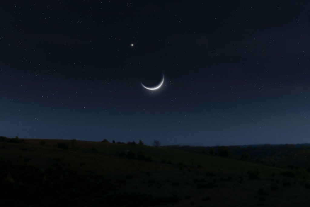 A imagem mostra um céu noturno estrelado com uma lua em fase nova bem fina, próxima ao horizonte. Abaixo, há uma paisagem escura de campo ou colina
