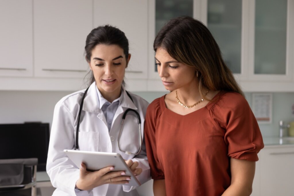 A imagem mostra uma médica com estetoscópio ao lado de uma paciente em um consultório. As duas observam um tablet, sugerindo que estão analisando informações médicas ou resultados de exames.