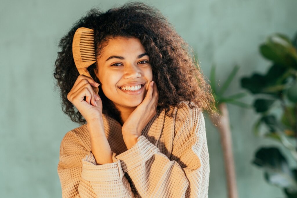 mulher de cabelo cacheado, roupão marrom, sorrindo e com pente perto do cabelo