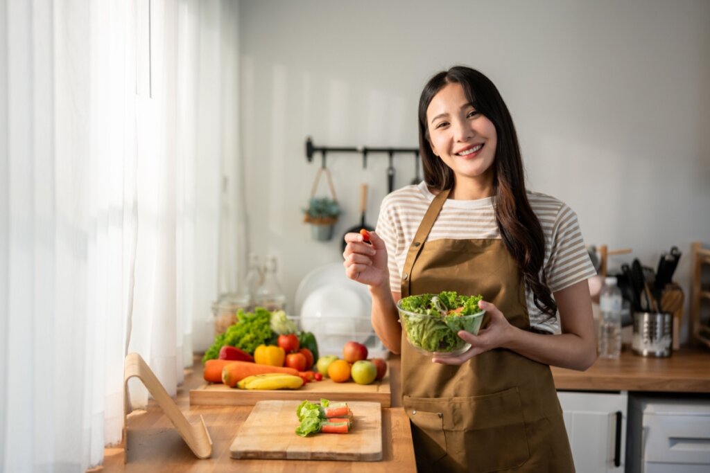 mulher de cabelo longo, avental marrom, sorrindo e segurando pote de vidro com salada verde. ela está ao lado de bancada com vegetais