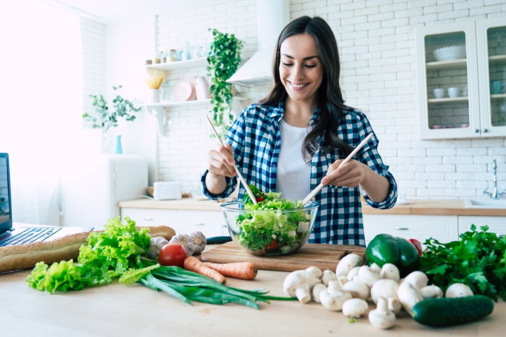 Mulher com cabelo preto solto, usando camiseta branca e camisa de botões xadrez em azul e branco preparando refeição saudável