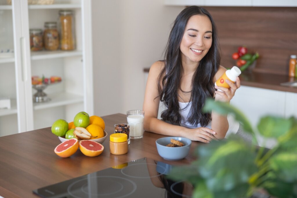 Uma mulher jovem sorridente, de cabelos escuros e longos, está sentada à mesa de uma cozinha iluminada. Ela segura um frasco de suplemento branco com rótulo laranja na mão esquerda, olhando para ele com atenção. À sua frente, sobre a mesa de madeira, há uma variedade de alimentos saudáveis: uma fruteira com maçãs, laranjas e peras, metades de uma toranja, um pote de pasta de amendoim, um copo de leite e uma tigela com biscoitos integrais. Ao fundo, vê-se uma estante branca com potes de mantimentos e parte da bancada da cozinha. A cena transmite uma atmosfera de bem-estar e cuidado com a saúde.