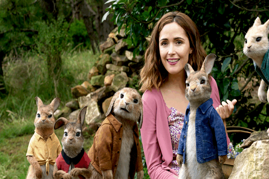 Mulher com cabelo curto, usando regata florida e casaco rosa, com 5 coelhos ao redor dela usando roupas humanas e ao fundo madeiras e a floresta