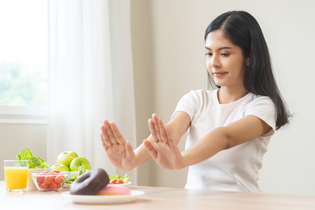 Mulher, sentada em frente a uma mesa de madeira com suco, frutas e donuts, recusando alimentos com as mãos