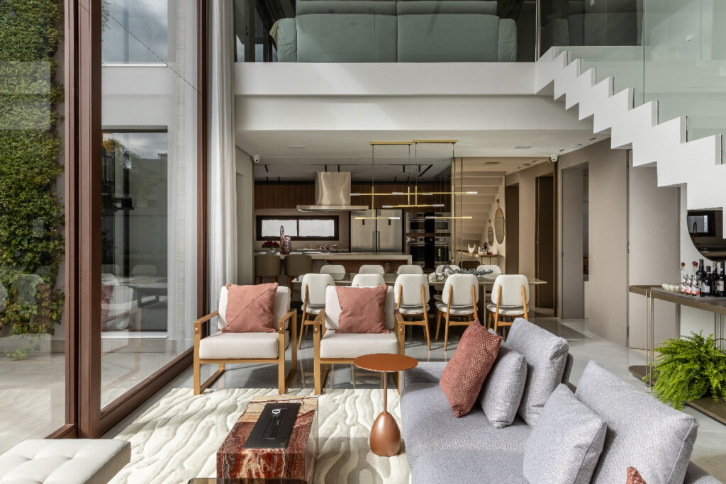 A imagem mostra uma sala de estar moderna integrada à cozinha e sala de jantar, com decoração elegante e minimalista. O ambiente é amplo e bem iluminado, com grandes janelas de vidro que permitem a entrada de luz natural. Há sofás confortáveis, poltronas e uma mesa de centro, além de uma escada que leva ao andar superior. Ao fundo, a cozinha planejada e a mesa de jantar completam o espaço, criando um cenário sofisticado e acolhedor.