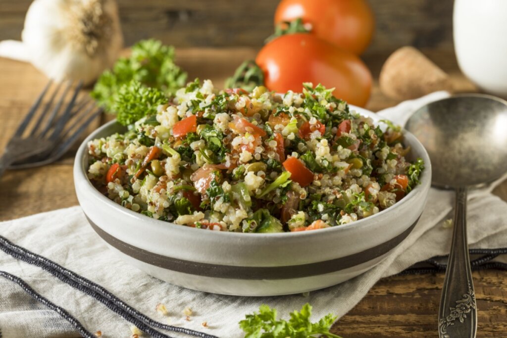 Salada de quinoa com legumes servida em tigela branca com listra marrom em cima de mesa de madeira