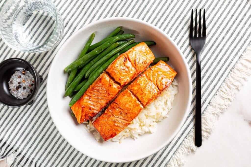 No centro, há um prato branco contendo dois pedaços de salmão grelhado, com a superfície dourada e levemente caramelizada, indicando que foram preparados na frigideira. O peixe está apoiado sobre uma porção de arroz branco soltinho. Ao lado, há vagens verdes cozidas