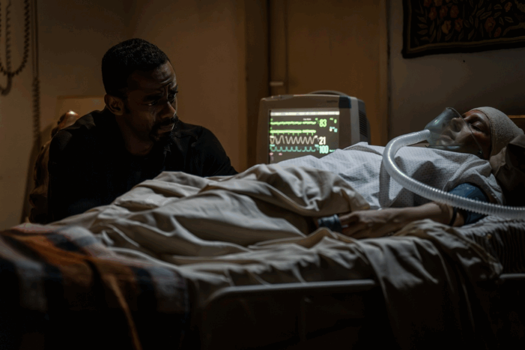 Homem com cabelo curto, barba, usando roupas pretas ao lado de mulher em cama de hospital ligada aos aparelhos 