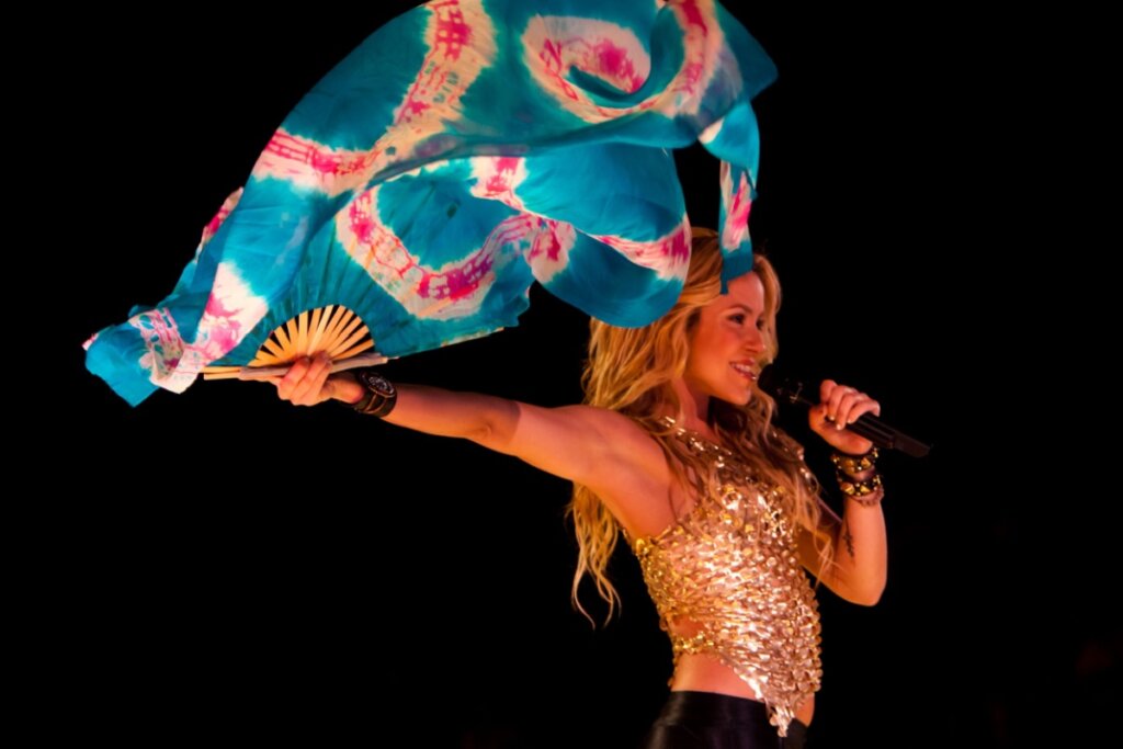 Imagem da cantora Shakira usando top dourado e calça preta e movimentando um leque com um tecido colorido em show