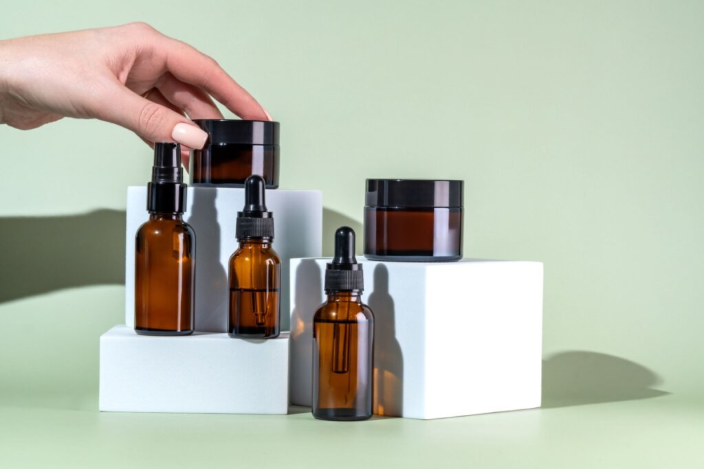 Produtos de skincare, com embalagem marrom, em cima de uma superfície verde com uma mão em cima de um deles