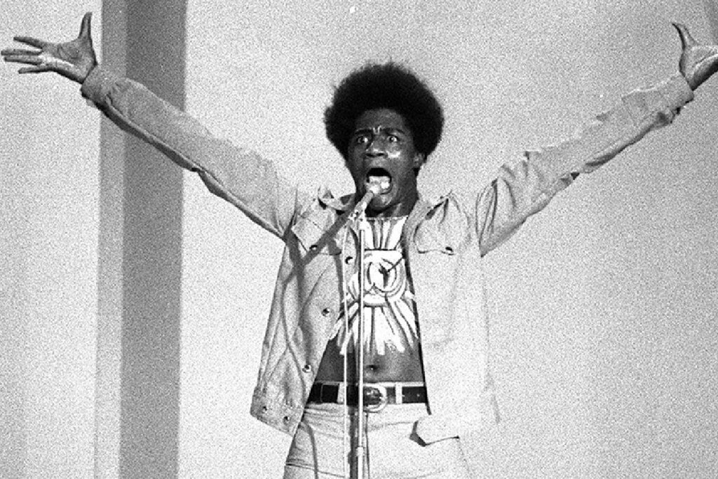 Fotografia em preto e branco de um homem negro com cabelo estilo black power, capturado em uma performance expressiva. Ele está com os braços estendidos para o alto e para os lados, com as mãos abertas e uma expressão facial intensa de canto ou grito. Ele veste uma jaqueta clara sobre uma camiseta com uma estampa circular e calças de cintura alta com cinto.