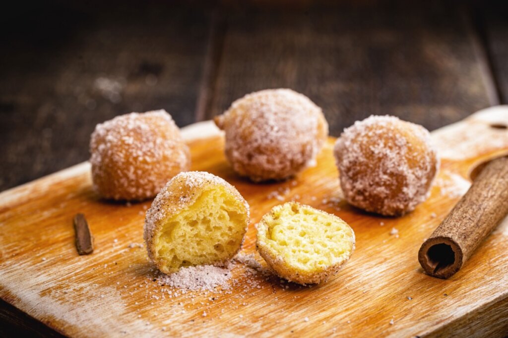 A imagem apresenta quatro bolinhos de chuva redondos dispostos sobre uma tábua de madeira rústica. Um dos bolinhos está partido ao meio em primeiro plano, revelando uma massa amarela clara, macia e aerada. Todos os bolinhos estão generosamente cobertos com uma mistura de açúcar e canela. Ao lado deles, há pedaços de canela em pau, e o fundo é composto por tons escuros de madeira, criando uma atmosfera acolhedora e rústica