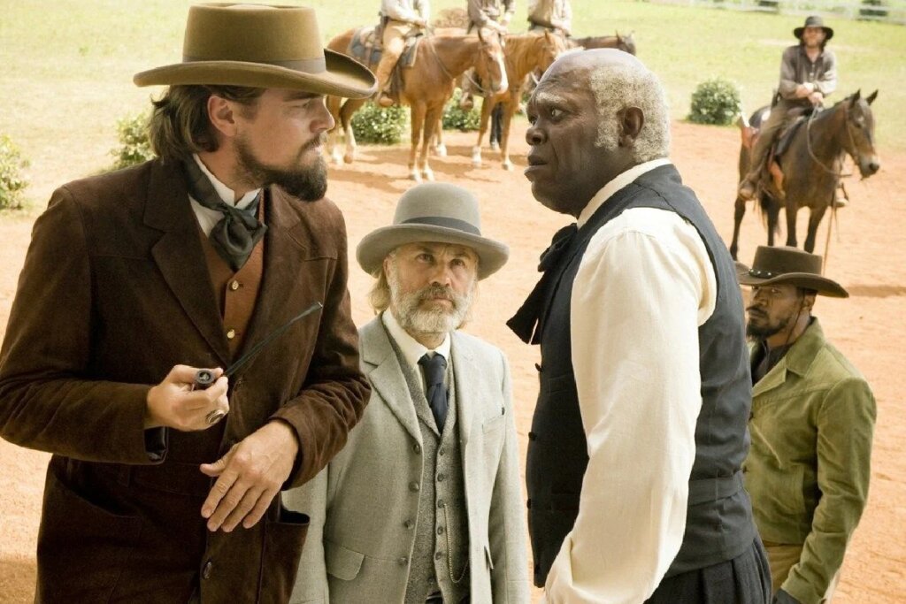 Em uma área externa ensolarada, Calvin Candie (Leonardo DiCaprio), vestindo paletó de veludo e chapéu marrom, encara Stephen (Samuel L. Jackson), um homem negro de cabelos brancos com expressão surpresa. Entre eles, o Dr. King Schultz observa a cena, enquanto Django aparece ao fundo à direita. No horizonte, homens montados a cavalo completam o cenário de uma fazenda do século XIX.