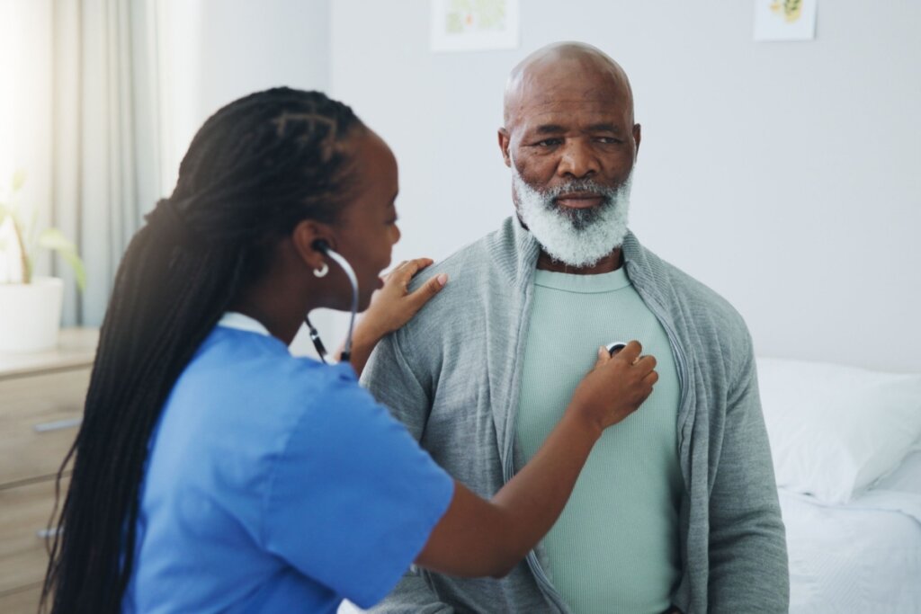 Uma enfermeira negra, de perfil e com longas tranças, utiliza um estetoscópio para auscultar o coração de um paciente idoso. O homem é negro, tem barba branca curta e veste um cardigã cinza sobre uma camiseta clara, mantendo uma expressão serena enquanto está sentado em um ambiente iluminado que sugere um quarto de hospital ou clínica. A enfermeira usa um uniforme azul e apoia gentilmente a mão direita no ombro do paciente.