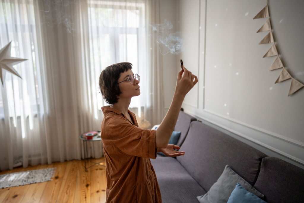 Uma jovem de pele clara, cabelos curtos escuros com franja e óculos de grau redondos está de perfil no centro de uma sala de estar. Ela segura uma lasca de madeira de Palo Santo acesa, observando a fumaça branca e densa que sobe em direção ao teto enquanto realiza uma defumação no ambiente. Ela veste um vestido midi de cor terracota. A sala possui piso de madeira clara, um sofá cinza com almofadas azuis e cinzas, cortinas brancas translúcidas na janela ao fundo e uma pequena bandeirinha de tecido pendurada na parede branca à direita, onde pequenos reflexos de luz pontilhada aparecem.