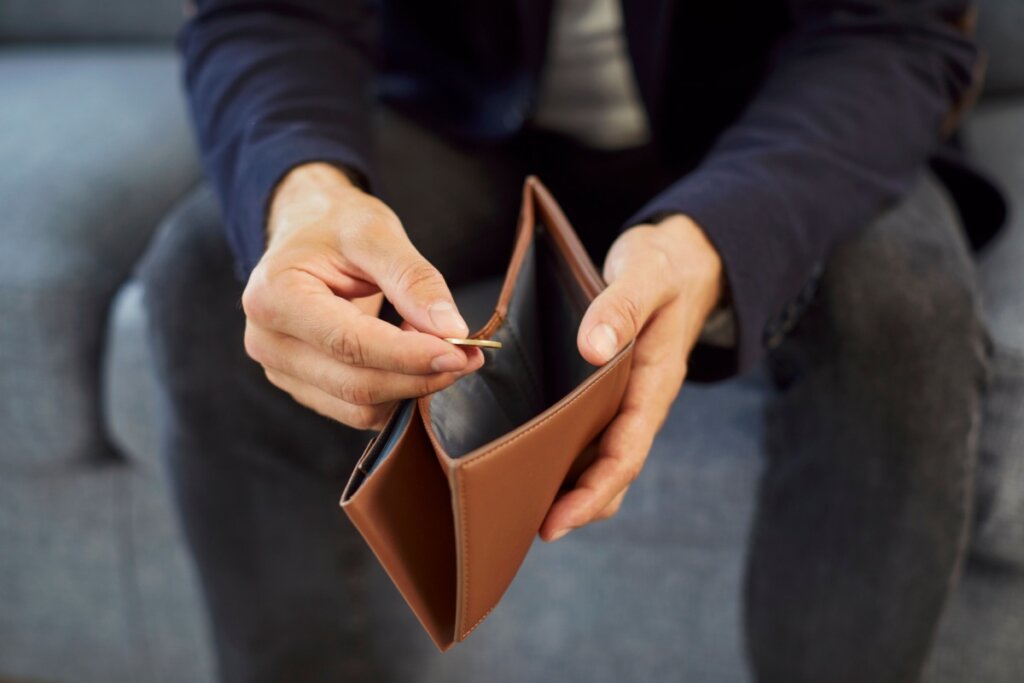 Um close nas mãos de uma pessoa sentada, que segura uma carteira de couro marrom aberta e vazia, exceto por uma única moeda de ouro que ela está retirando. O resto da pessoa e o ambiente estão desfocados.