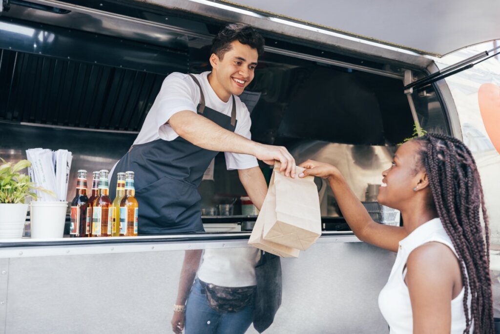 Uma mulher negra com tranças longas sorri ao receber duas sacolas de papel das mãos de um atendente jovem dentro de um food truck. O atendente, um homem jovem de pele clara e cabelos escuros, veste camiseta branca e avental cinza escuro, estendendo o braço sorridente para entregar o pedido. No balcão de metal do veículo, há garrafas de vidro com bebidas coloridas e um pequeno vaso de planta. A cena tem iluminação natural diurna e transmite simpatia no atendimento.