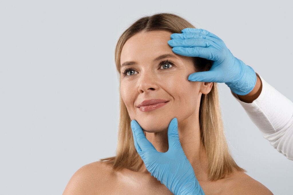 Rosto de uma mulher loira, sorrindo levemente enquanto é examinada. Duas mãos usando luvas cirúrgicas azuis tocam suavemente sua face — uma na têmpora e outra no queixo —, simulando uma avaliação para procedimentos estéticos ou cirurgia plástica. O fundo é cinza claro e neutro.