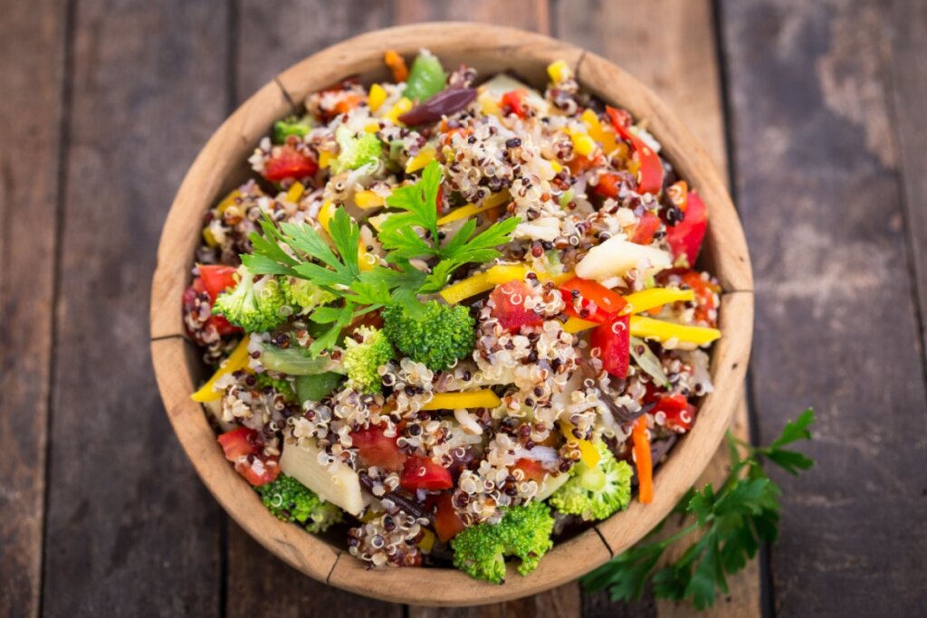 Quinoa com brócolis, pimentões coloridos, legumes variados e folhas frescas servida em tigela de madeira