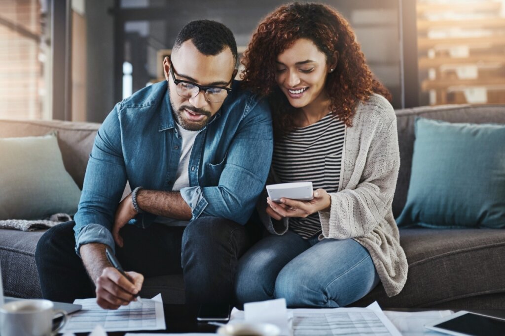 Casal sentado em um sofá cinza, analisando documentos financeiros. O homem escreve em papéis com uma caneta, enquanto a mulher usa uma calculadora e sorri. Há planilhas, celular, tablet e uma xícara de café sobre a mesa. Ambiente de sala de estar iluminado por luz natural.