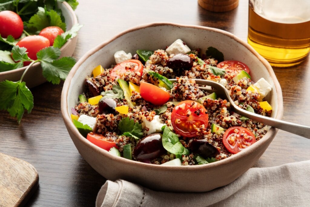 Uma tigela de cerâmica rústica contém uma salada mediterrânea colorida composta por uma base de quinoa branca e vermelha. A mistura inclui tomates cereja cortados ao meio, cubos de queijo feta branco, azeitonas pretas inteiras, pedaços de pepino, pimentão amarelo picado e folhas frescas de coentro.