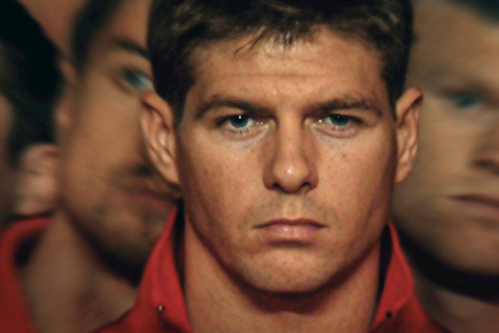 Um close-up dramático e focado no rosto do ex-jogador de futebol Steven Gerrard. Ele tem uma expressão séria, lábios cerrados e olhos azuis fixos diretamente para a câmera. Ele veste uma jaqueta esportiva vermelha com gola levantada. O fundo está desfocado com um efeito de sobreposição que duplica de forma suave as laterais de seu próprio rosto, criando uma atmosfera intensa e psicológica.
