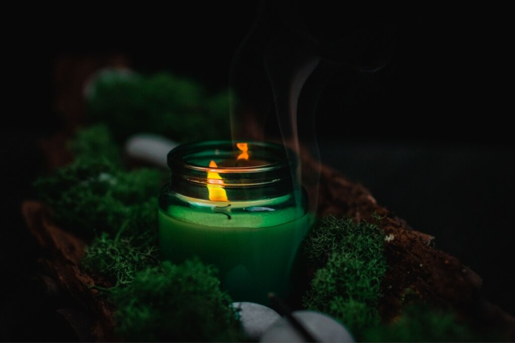 Uma vela verde acesa em um pote de vidro verde esmeralda, posicionada no centro de um arranjo rústico. A chama amarela e vibrante emite uma fumaça leve e fluida que sobe em direção ao topo da imagem. O pote está cercado por musgo verde denso e pedras pequenas e claras, tudo disposto sobre uma base de madeira escura com textura de casca de árvore. O fundo é preto, criando um contraste dramático que destaca a iluminação suave da vela e os tons de verde.