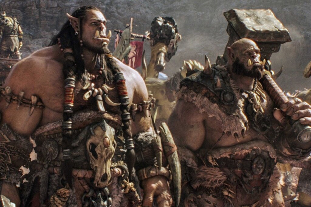 Uma cena de plano médio mostrando dois orcs da franquia Warcraft em um ambiente árido e rochoso. À esquerda, um orc de pele marrom com longas tranças adornadas por anéis metálicos e presas proeminentes que saem da mandíbula inferior. Ele usa colares de ossos e uma armadura de couro com um crânio animal na cintura. À direita, outro orc de barba escura segura um enorme martelo de pedra sobre o ombro. Ao fundo, estandartes de guerra e outros membros da horda completam o cenário épico.