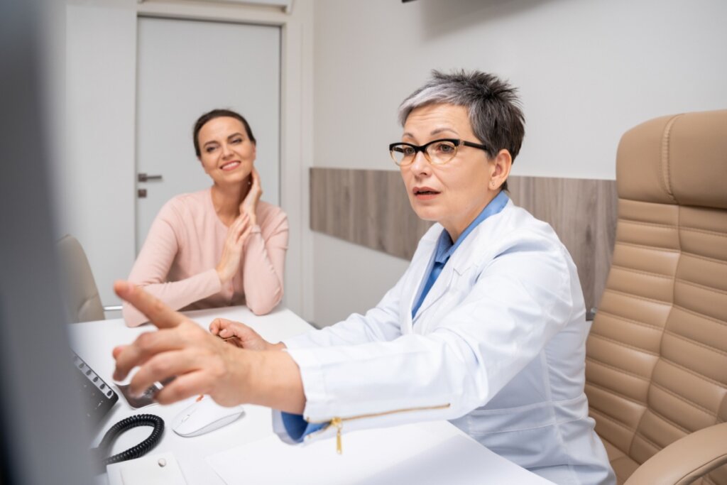 Médica mostrando algo para uma mulher em tela de computador. Ambas sentadas em um consultório