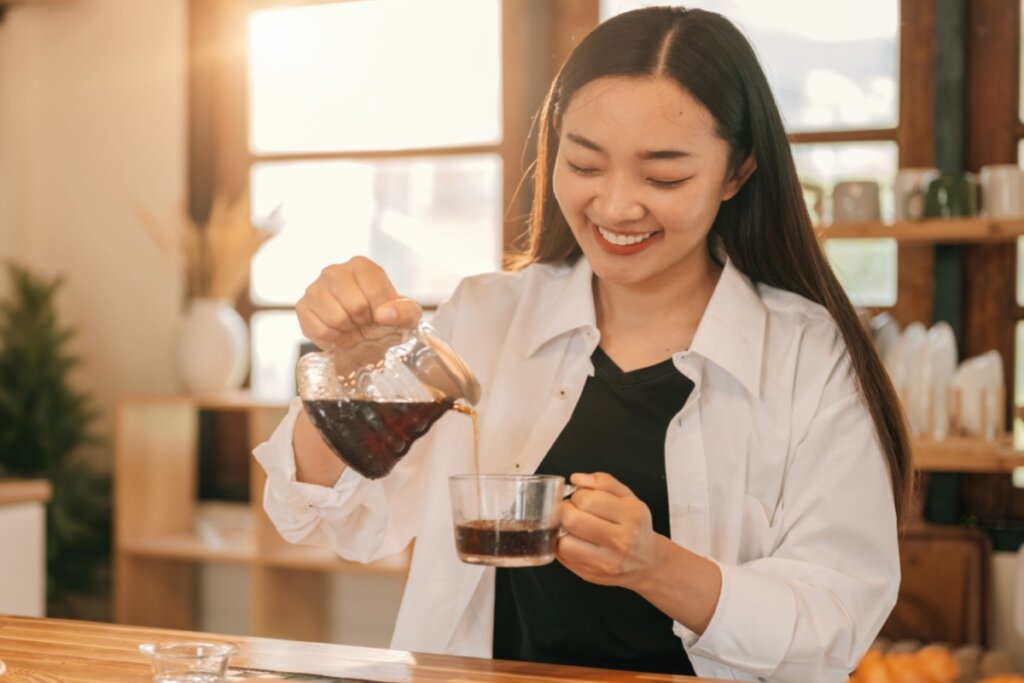 Mulher preparando café ao despejar a bebida em uma xícara, enquanto sorri