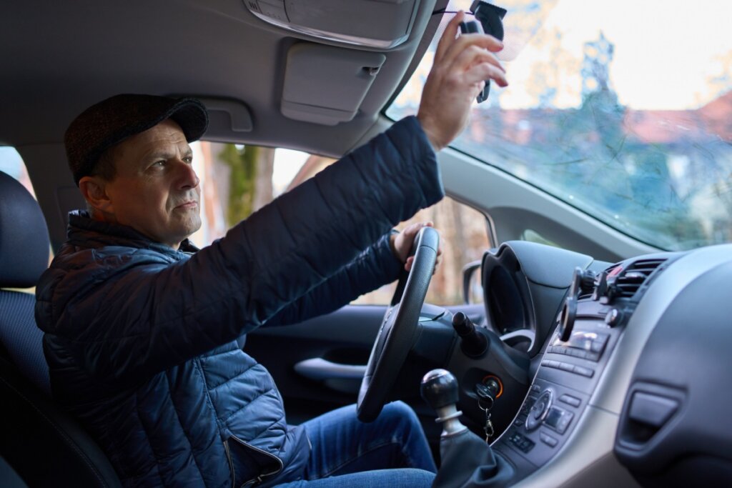 Homem sentado em um carro, usando casaco e boina preta, checando o retrovisor