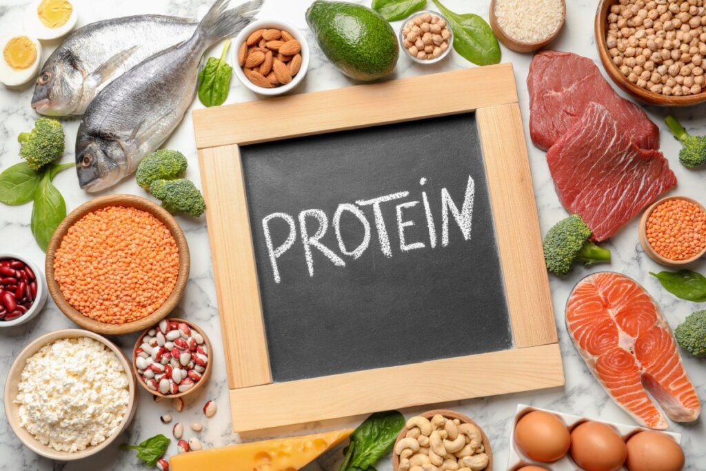 Alimentos ricos em proteína como carne, ovos, peixe, lentilha em cima de bancada de pedra branca com uma lousa escrito proteína em inglês