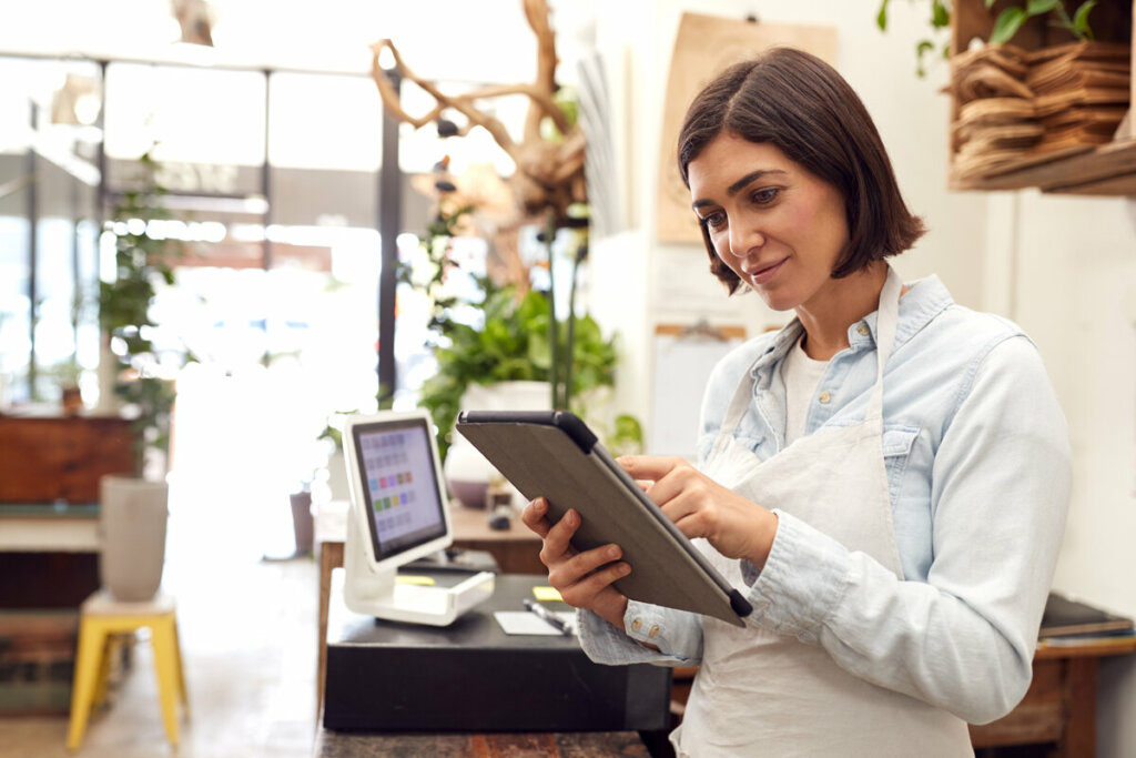 Uma mulher de cabelos curtos e castanhos, vestindo uma camisa jeans clara e um avental branco, opera um tablet em uma loja ou cafeteria. Ela está atrás de um balcão de madeira onde se vê um sistema de caixa branco ao fundo. O ambiente é iluminado e decorado com plantas, sugerindo um comércio moderno e acolhedor. Ela mantém uma expressão focada e um leve sorriso enquanto interage com a tela.