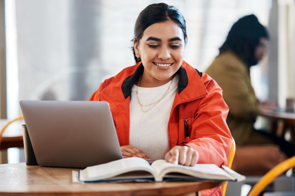 menina de jaqueta laranja, sentada, sorrindo e com mão em notebook e livro em mesa