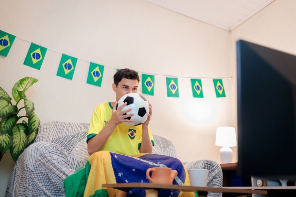 Homem sentado em um sofá, segurando uma bola de futebol em frente ao rosto, enquanto assiste ao jogo